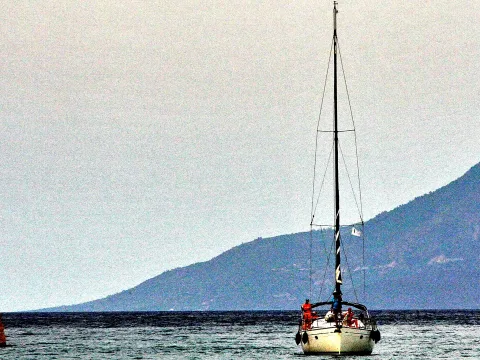Ein weißes Segelboot mit hohem Mast fährt auf ruhigem Meer, im Hintergrund zieht sich ein bewaldeter Berg zur Küste.