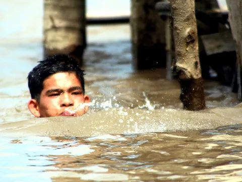 Junger Mann schwimmt im braunen Wasser, nur Kopf und Schultern sind sichtbar, vor seinem Mund spritzt Wasser auf. Im Hintergrund stehen hölzerne Pfähle.