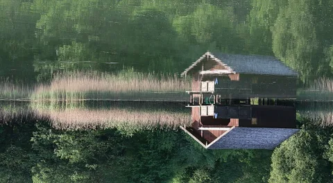 Eine dunkle Holzhütte mit Satteldach steht am Seeufer, davor wächst hohes Schilf, im ruhigen Wasser spiegeln sich grüne Bäume.