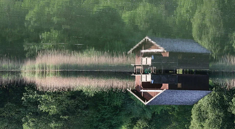 Eine dunkle Holzhütte mit Satteldach steht am Seeufer, davor wächst hohes Schilf, im ruhigen Wasser spiegeln sich grüne Bäume.