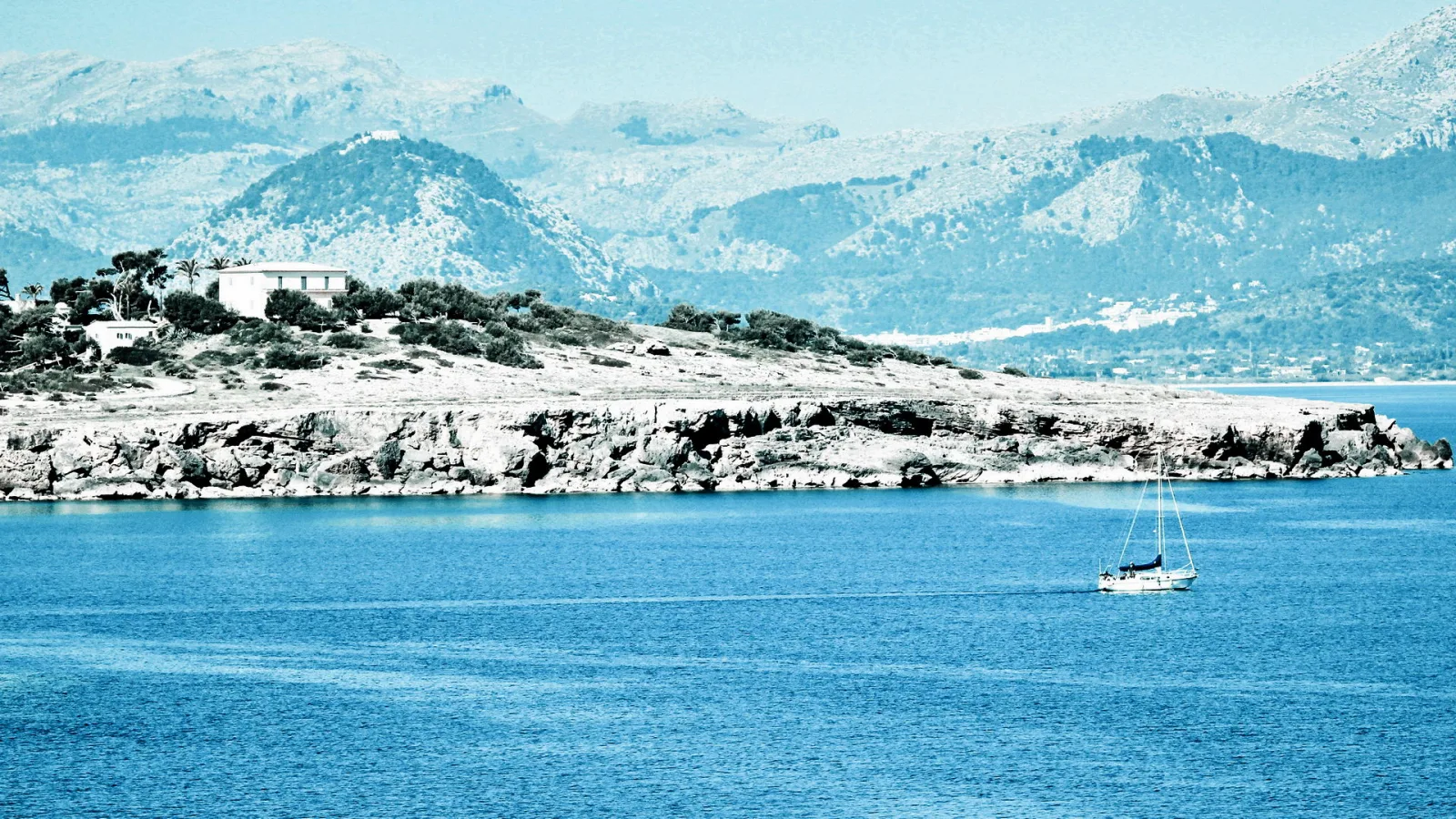 Ein weißes Segelboot fährt auf ruhigem blauem Meer. Links ragt eine felsige Küste mit einem weißen Haus und Bäumen ins Wasser, dahinter erheben sich blassblaue Berge unter hellem Himmel.