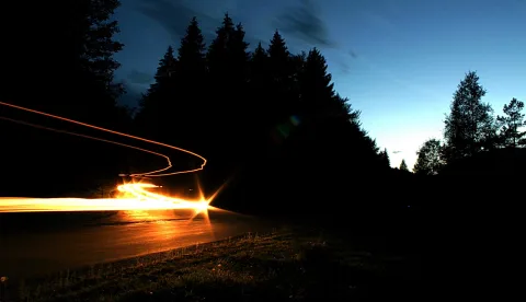 Eine Landstraße am Abend mit einer langen, gebogenen Lichtspur von Scheinwerfern vor einer dunklen Silhouette aus Nadelbäumen und einem blauen Himmel.