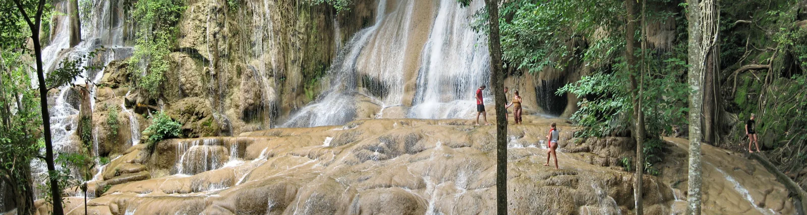 Mehrstufiger Wasserfall stürzt über beige Kalksteinterrassen, davor stehen vier Menschen zwischen Bäumen und dichtem Grün.
