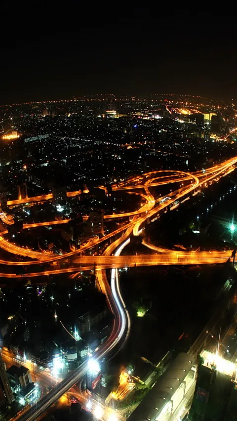 Mehrstöckiges Autobahnkreuz bei Nacht, orangefarbene Lichtspuren von Fahrzeugen auf den Fahrbahnen, dahinter eine beleuchtete Stadt unter dunklem Himmel.