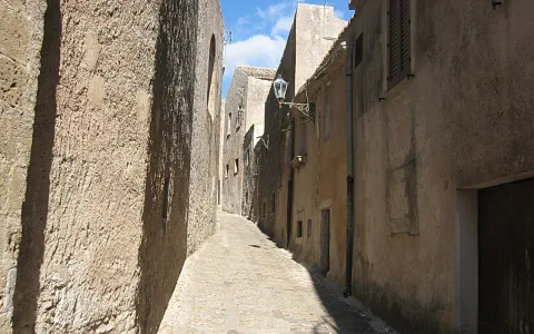Schmale Kopfsteinpflastergasse zwischen sandfarbenen Steinhäusern, links eine Mauer im Schatten, oben ein blauer Himmel mit Wolken und eine Laterne an der rechten Hauswand.