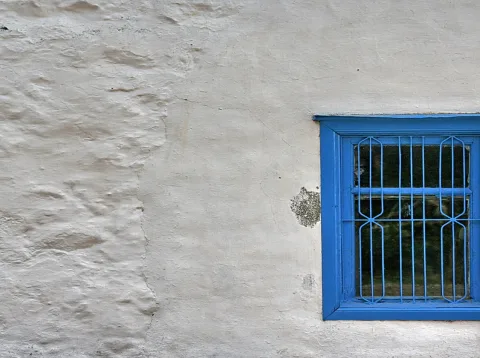 Rau verputzte, weiß gekalkte Hauswand mit einem blau lackierten Fenster rechts, vor dem ein schmiedeeisernes Gitter angebracht ist.