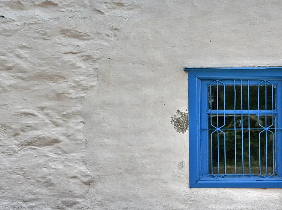 Rau verputzte, weiß gekalkte Hauswand mit einem blau lackierten Fenster rechts, vor dem ein schmiedeeisernes Gitter angebracht ist.