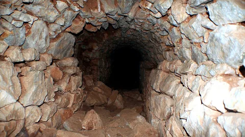 Enger Tunnel mit unregelmäßig geschichteten Steinwänden und Geröll am Boden, am Ende eine dunkle Öffnung.