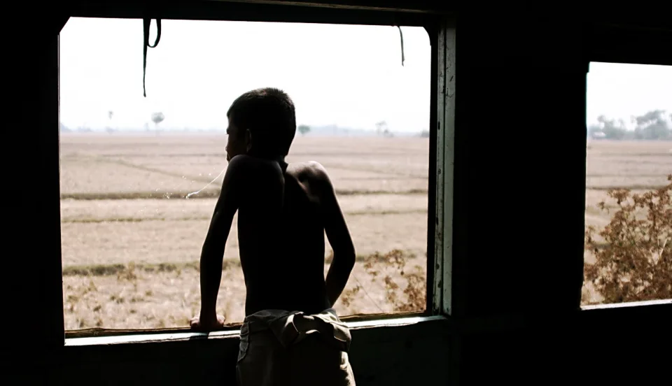 Silhouette eines Jungen mit nacktem Oberkörper, der aus einem Zugfenster auf trockene Felder blickt.