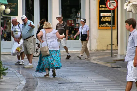 Mehrere Touristen gehen über eine gepflasterte Gasse, im Vordergrund eine Frau in weißer Bluse und blau geblümtem Rock, dahinter Männer mit Hüten und Kappen sowie ein junger Mann am rechten Bildrand.