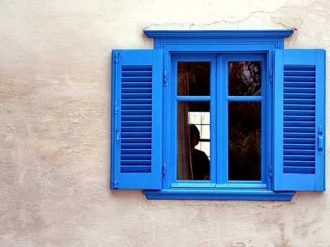 Blau lackierte Fensterläden rahmen ein Fenster in einer hellen Putzwand, im Fenster ist eine dunkle Silhouette einer Person zu sehen.