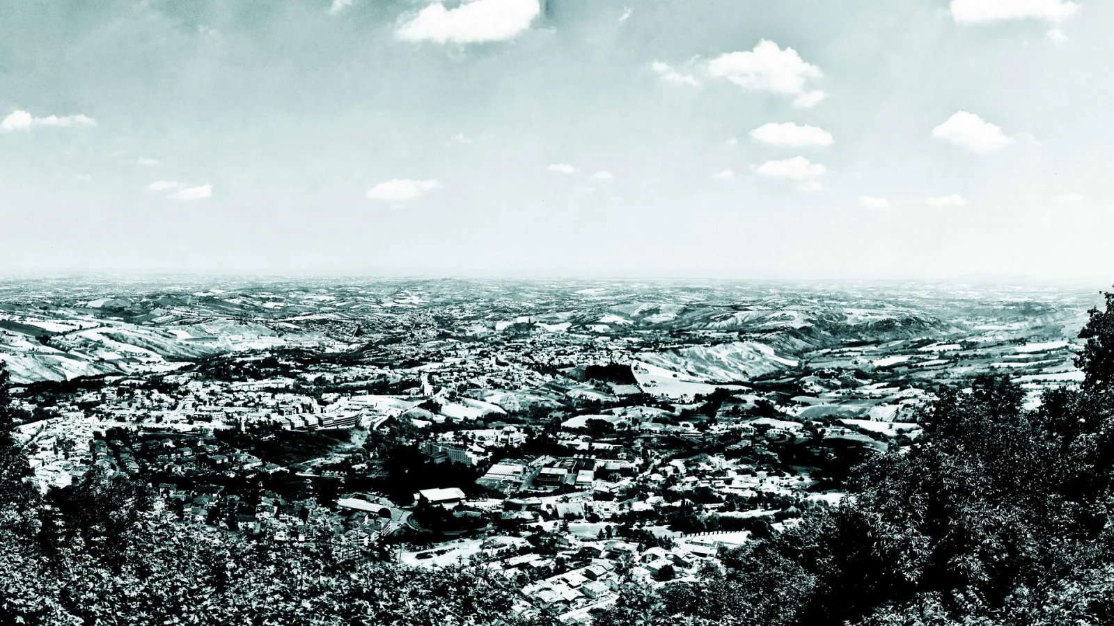 Schwarz-weiße Panoramaaufnahme einer Stadt in hügeliger Landschaft, im Vordergrund dichte Bäume, darüber ein weiter Himmel mit Wolken.