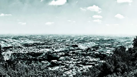 Schwarz-weiße Panoramaaufnahme einer Stadt in hügeliger Landschaft, im Vordergrund dichte Bäume, darüber ein weiter Himmel mit Wolken.