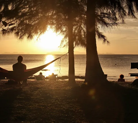 Silhouette eines Mannes in einer Hängematte zwischen Bäumen am Strand, im Hintergrund geht die Sonne über dem Meer unter.