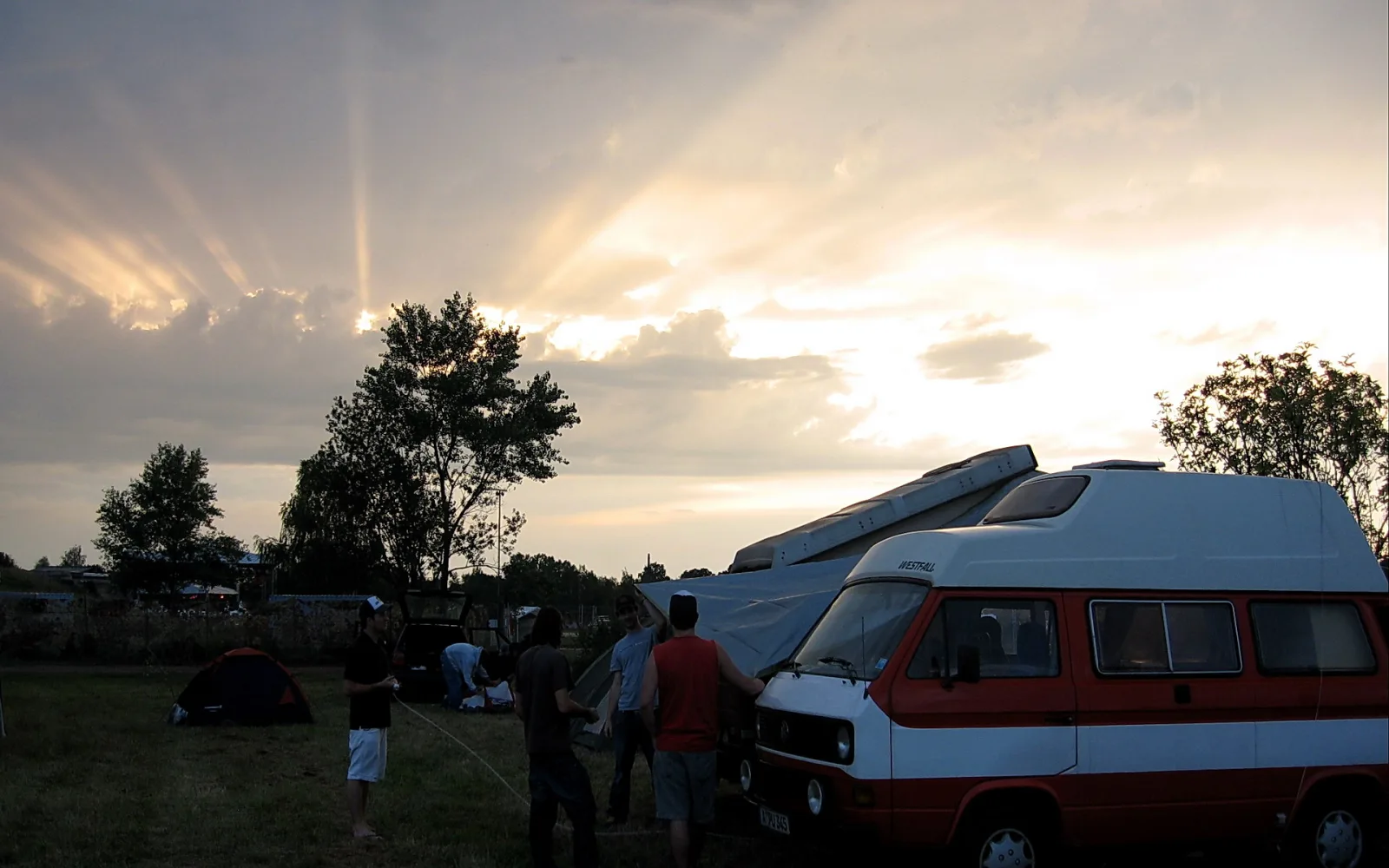 Menschen bauen bei Abendlicht ein Zelt auf, daneben steht ein rot-weißer Campingbus, darüber brechen Sonnenstrahlen durch die Wolken.