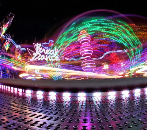 Nachtaufnahme eines Karussells auf dem Jahrmarkt mit bunten Lichtspuren, im Hintergrund die beleuchtete Schrift Break Dance.