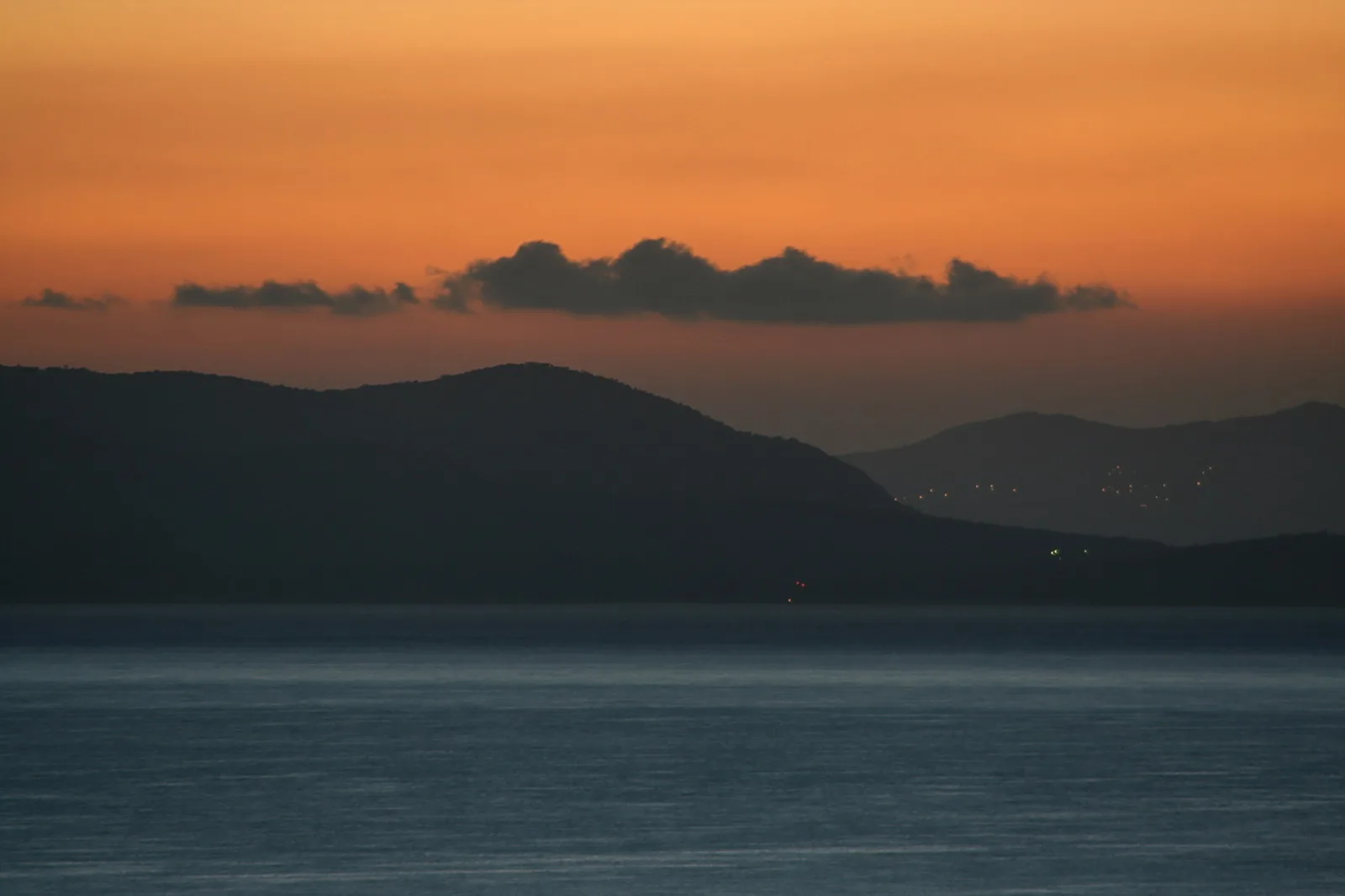 Dunkle Bergkette vor einer glatten Meeresfläche unter orangefarbenem Abendhimmel mit einzelnen Wolkenbändern