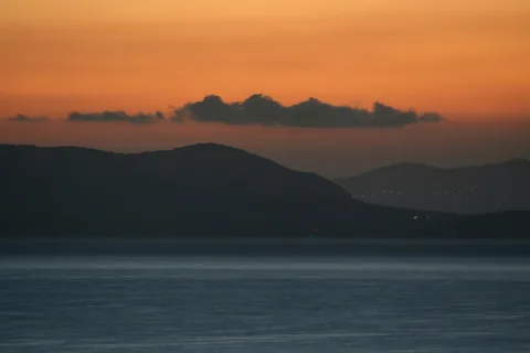 Dunkle Bergkette vor einer glatten Meeresfläche unter orangefarbenem Abendhimmel mit einzelnen Wolkenbändern