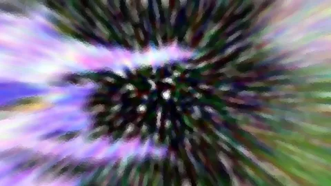 Abstrakte Aufnahme mit radialem Zoomeffekt. Von einem dunklen, körnigen Zentrum strahlen violette, grüne und weiße Schlieren nach außen.