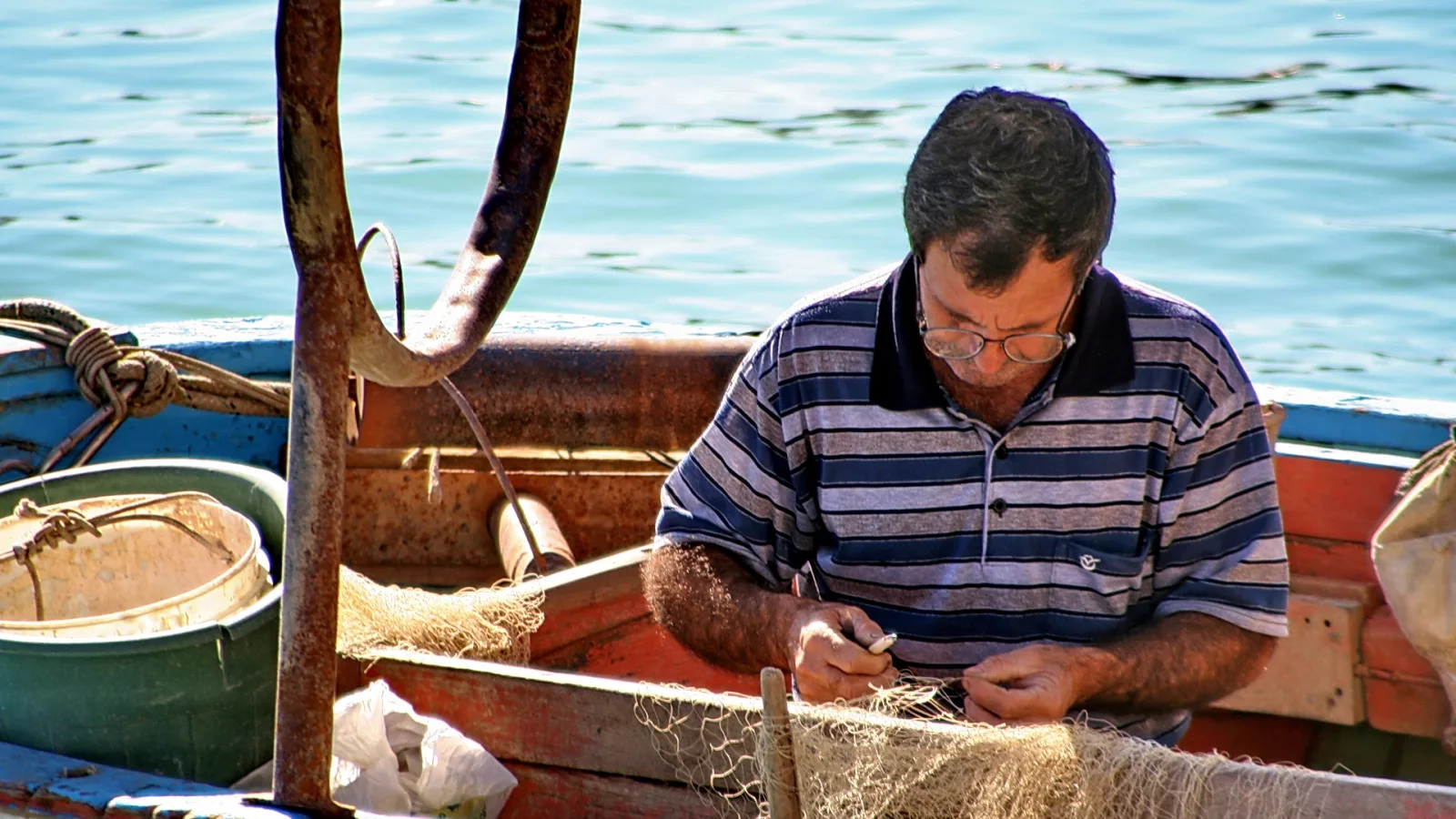 Ein Mann mit Brille und gestreiftem Polohemd sitzt in einem Fischerboot und flickt ein Fischernetz, im Hintergrund liegt blaues Meerwasser.