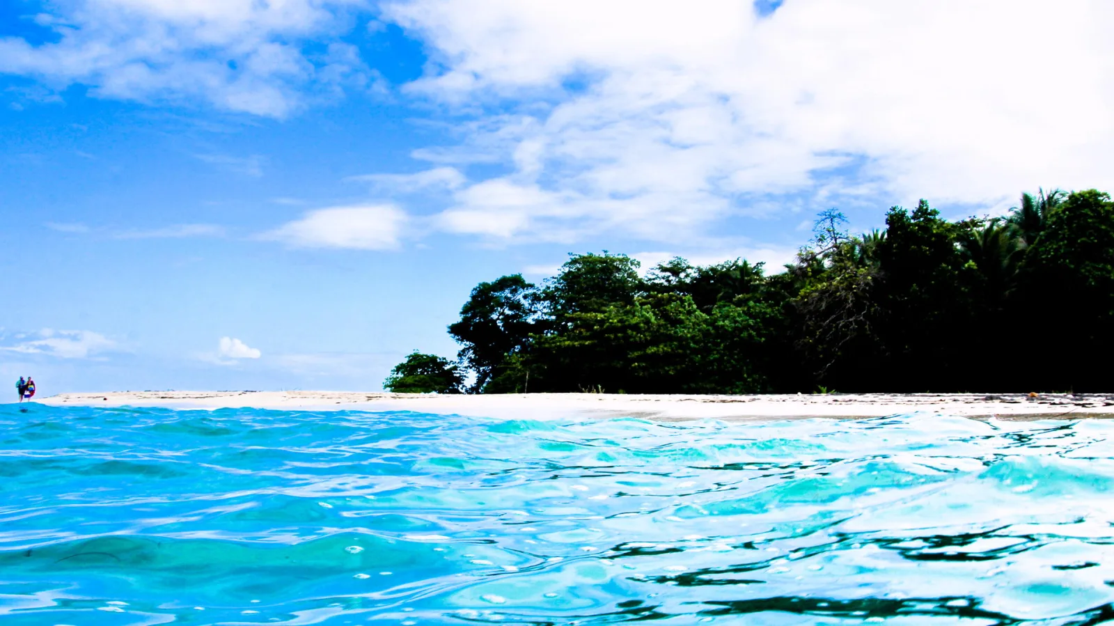 Türkisfarbenes, leicht bewegtes Meer im Vordergrund, dahinter ein heller Sandstrand und dichter grüner Küstenwald unter blauem Himmel mit weißen Wolken. Links im Hintergrund steht eine winzige Person im Wasser.