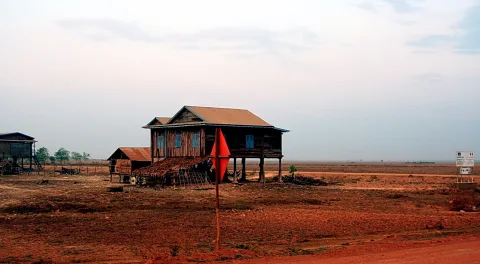 Ein hölzernes Haus auf Stelzen steht in einer flachen, trockenen, rötlich-braunen Ebene unter hellem, dunstigem Himmel. Links steht eine kleine Hütte, rechts ein Schild, und vorne ragt eine rote Fahne auf.
