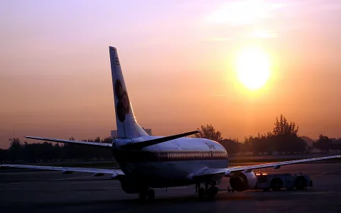 Ein Passagierflugzeug steht von hinten gesehen auf dem Vorfeld, rechts davon zieht ein Schlepper, im Hintergrund stehen Bäume und die tiefstehende Sonne färbt den Dunst orange.