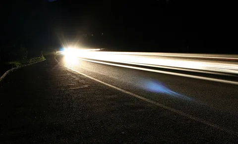 Langzeitbelichtung einer kurvigen Straße bei Nacht. Helle Scheinwerferspuren ziehen von links nach rechts über den Asphalt, links verläuft eine Leitplanke.