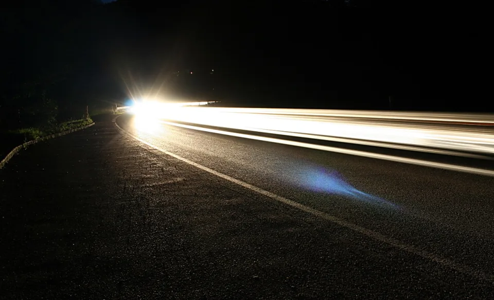 Langzeitbelichtung einer kurvigen Straße bei Nacht. Helle Scheinwerferspuren ziehen von links nach rechts über den Asphalt, links verläuft eine Leitplanke.