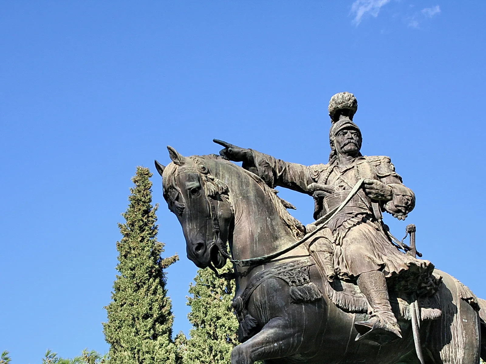 Bronzestatue eines Reiters auf einem Pferd, der den rechten Arm nach vorn ausstreckt, mit Fellmütze und Uniform, vor blauem Himmel und Zypressen.