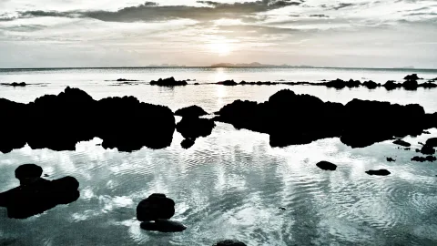 Dunkle Felsen ragen aus flachem Wasser, im Hintergrund geht die Sonne hinter einer Wolkendecke über dem Meer unter.