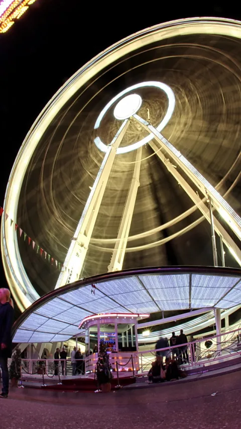 Ein beleuchtetes Riesenrad vor schwarzem Nachthimmel, dessen Lichtkreise durch die lange Belichtung zu Spuren verwischen. Darunter eine überdachte Plattform mit Menschen und einem kleinen geschmückten Weihnachtsbaum.