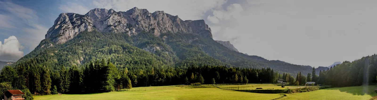 Breites Panorama einer schroffen, grau-hellen Felswand, die über dunkelgrünen Nadelwäldern und einer sonnigen grünen Wiese aufragt, links ein rotbraunes Holzhaus am Waldrand, der Himmel blau mit weißen Wolken.