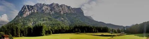 Breites Panorama einer schroffen, grau-hellen Felswand, die über dunkelgrünen Nadelwäldern und einer sonnigen grünen Wiese aufragt, links ein rotbraunes Holzhaus am Waldrand, der Himmel blau mit weißen Wolken.