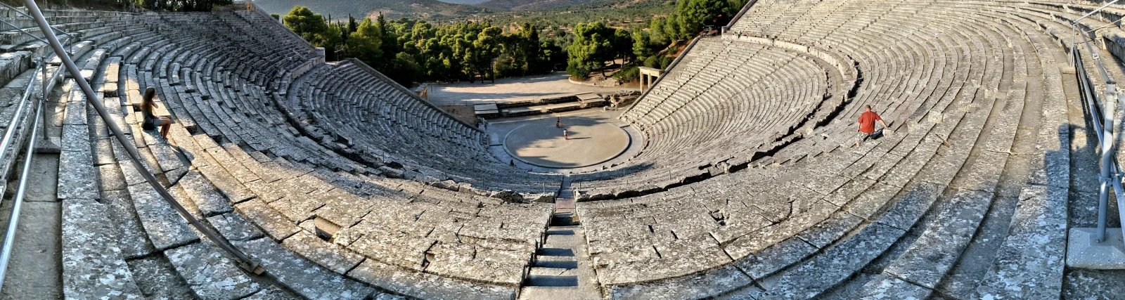 Panoramablick auf die steinernen Sitzreihen des antiken Theaters von Epidaurus, in der Mitte die runde Spielfläche, rechts eine rot gekleidete Person auf den Stufen.