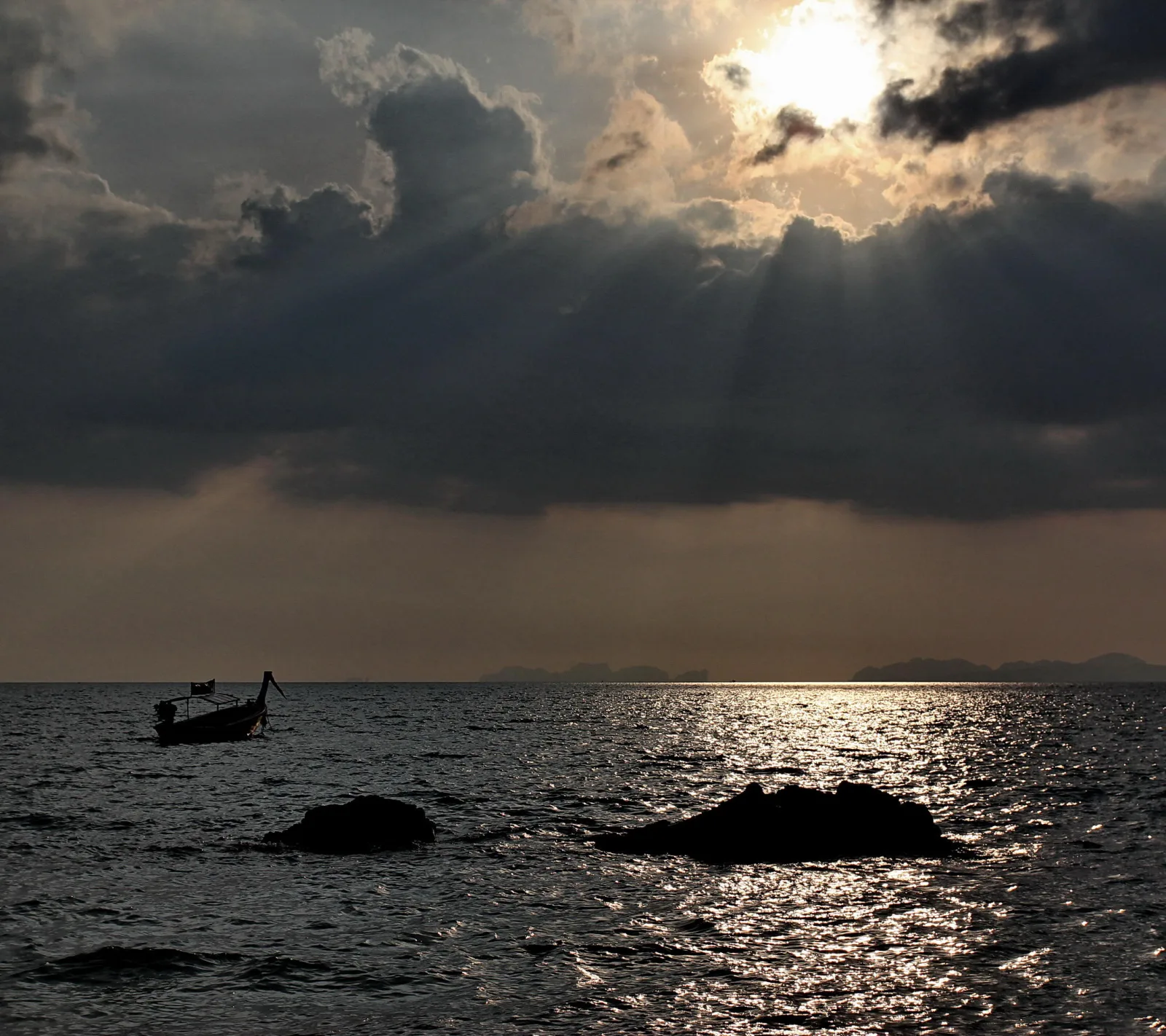 Silhouette eines langen Holzboots auf dunklem Meer, links im Bild, darüber Wolken mit Sonnenstrahlen, im Vordergrund zwei Felsen im Wasser, rechts glitzert die Sonne auf den Wellen.