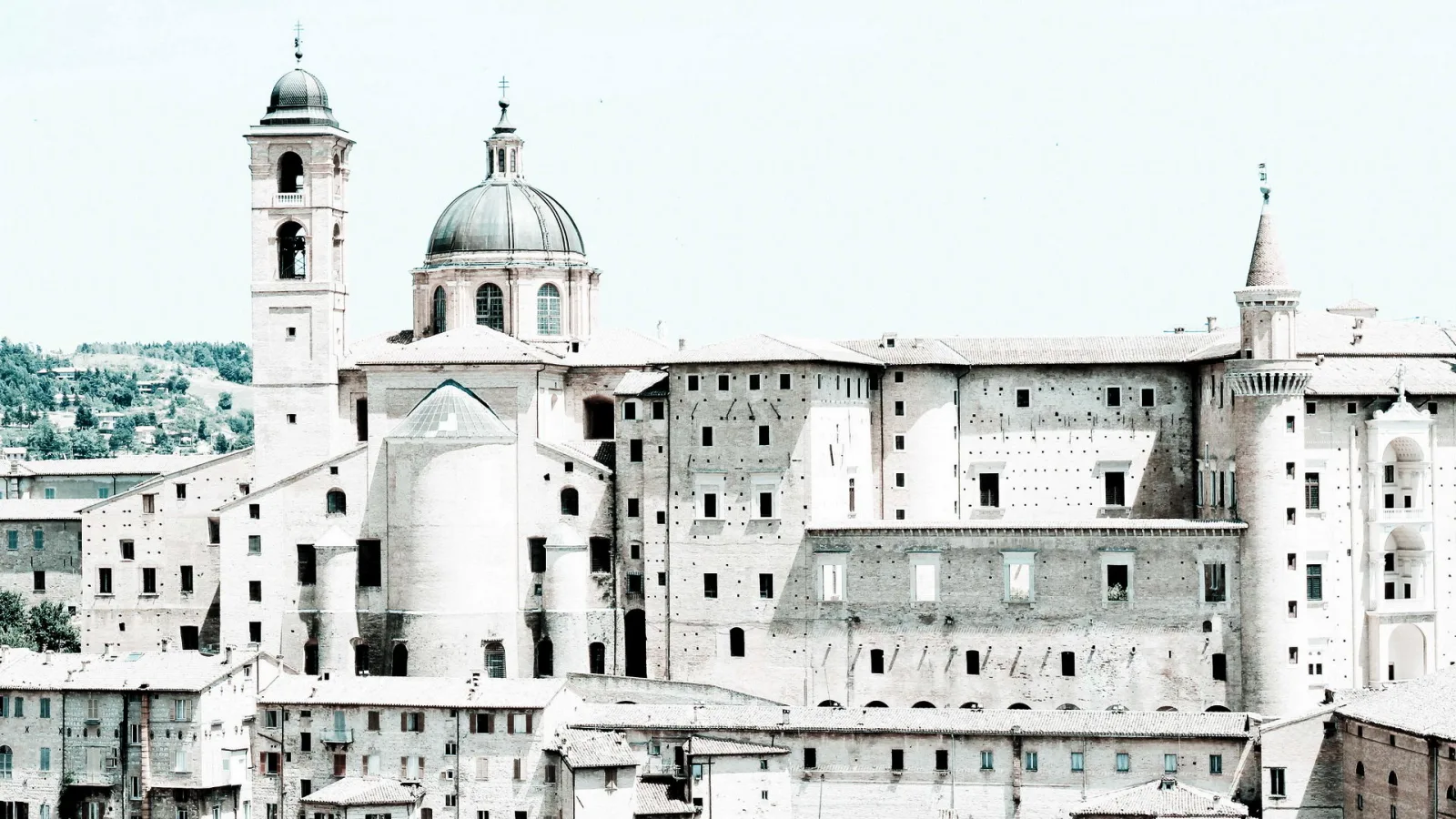 Blick auf die Altstadt von Urbino mit Domkuppel, Glockenturm und einem großen Renaissancepalast aus hellem Backstein unter blassem Himmel.