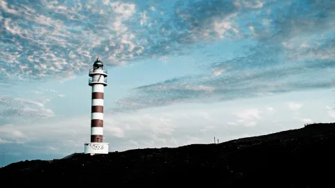 Rot-weiß gestreifter Leuchtturm auf einem dunklen Hügel unter einem blauen, wolkigen Himmel.