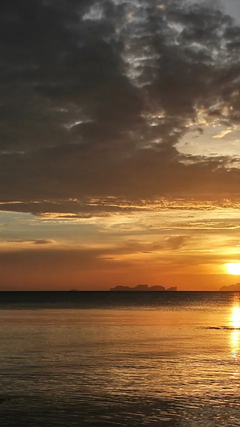 Sonnenuntergang über einer ruhigen Meeresfläche, die Sonne sitzt rechts am Horizont, darüber dichte dunkle Wolken, im Hintergrund die Silhouette kleiner Inseln.