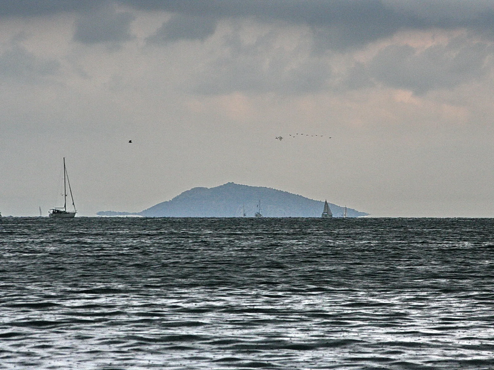 Eine dunstige Insel am Horizont, davor bewegtes graues Meer mit mehreren Segelbooten unter bedecktem Himmel.