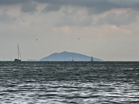 Eine dunstige Insel am Horizont, davor bewegtes graues Meer mit mehreren Segelbooten unter bedecktem Himmel.