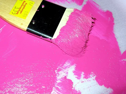 Flacher Pinsel mit pinker Farbe auf einer großflächig bemalten Fläche, im Griff ein gelbes Preisschild mit handschriftlicher Zahl.