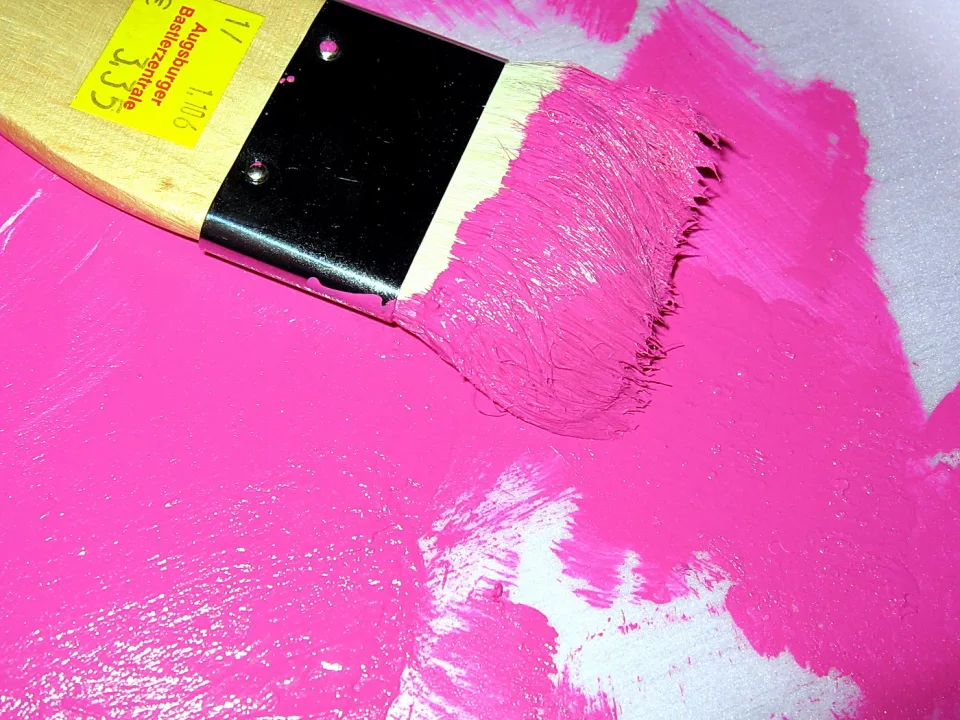 Flacher Pinsel mit pinker Farbe auf einer großflächig bemalten Fläche, im Griff ein gelbes Preisschild mit handschriftlicher Zahl.