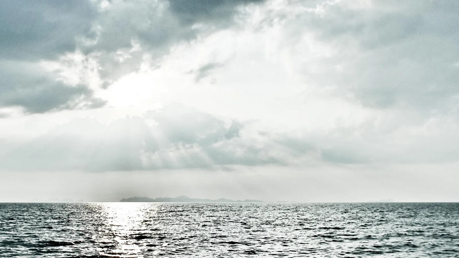 Graues Meer unter bewölktem Himmel, Sonnenstrahlen brechen durch die Wolken, am Horizont liegt eine flache Insel.