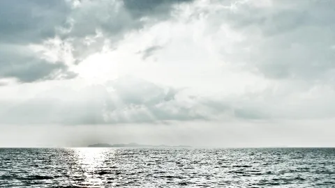 Graues Meer unter bewölktem Himmel, Sonnenstrahlen brechen durch die Wolken, am Horizont liegt eine flache Insel.