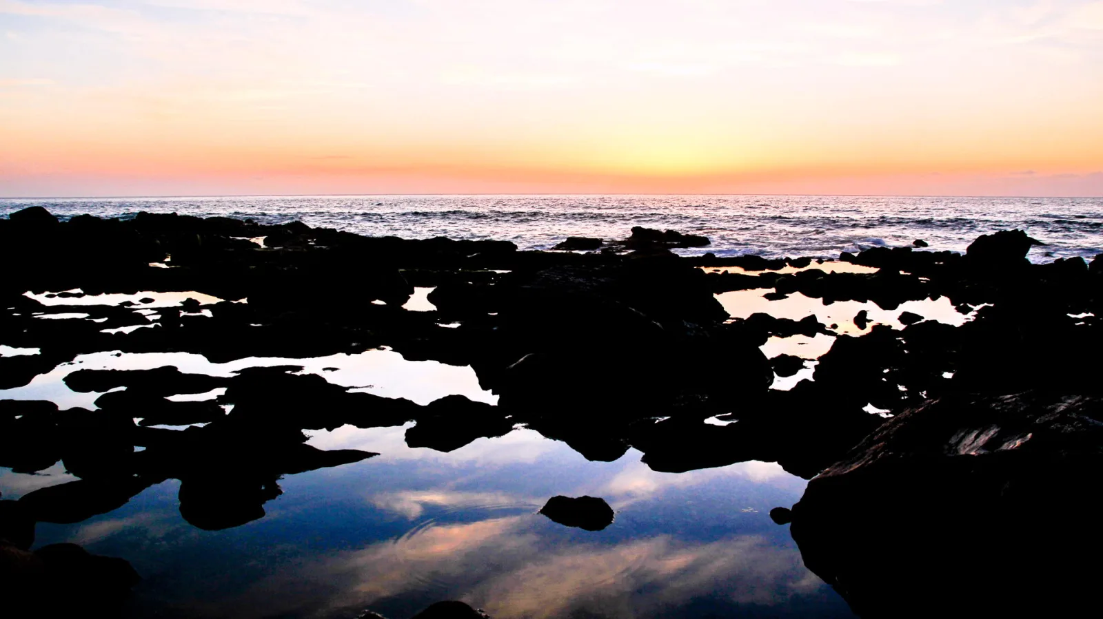 Dunkle Felsen mit Wasserpfützen vor dem Meer bei Sonnenuntergang, der Himmel leuchtet orange und gelb.