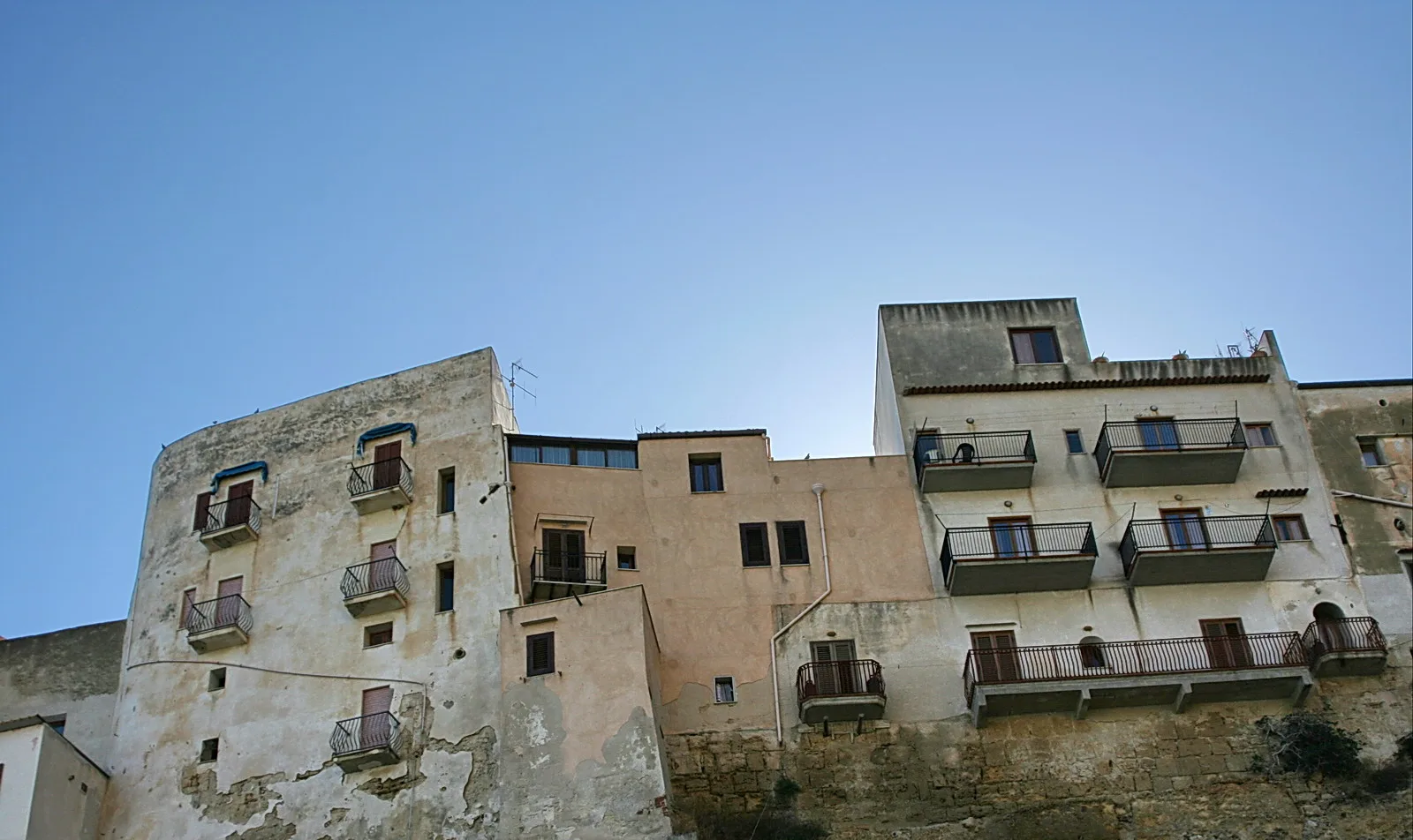 Verwitterte mehrstöckige Häuserfassaden mit Balkonen und Fensterläden vor klarem blauem Himmel an einem Felshang.