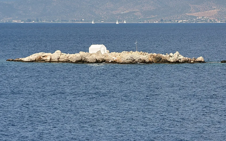 Kleine weiße Kapelle auf einem felsigen Inselchen im blauen Meer, dahinter Hügel und zwei Segelboote am Horizont.