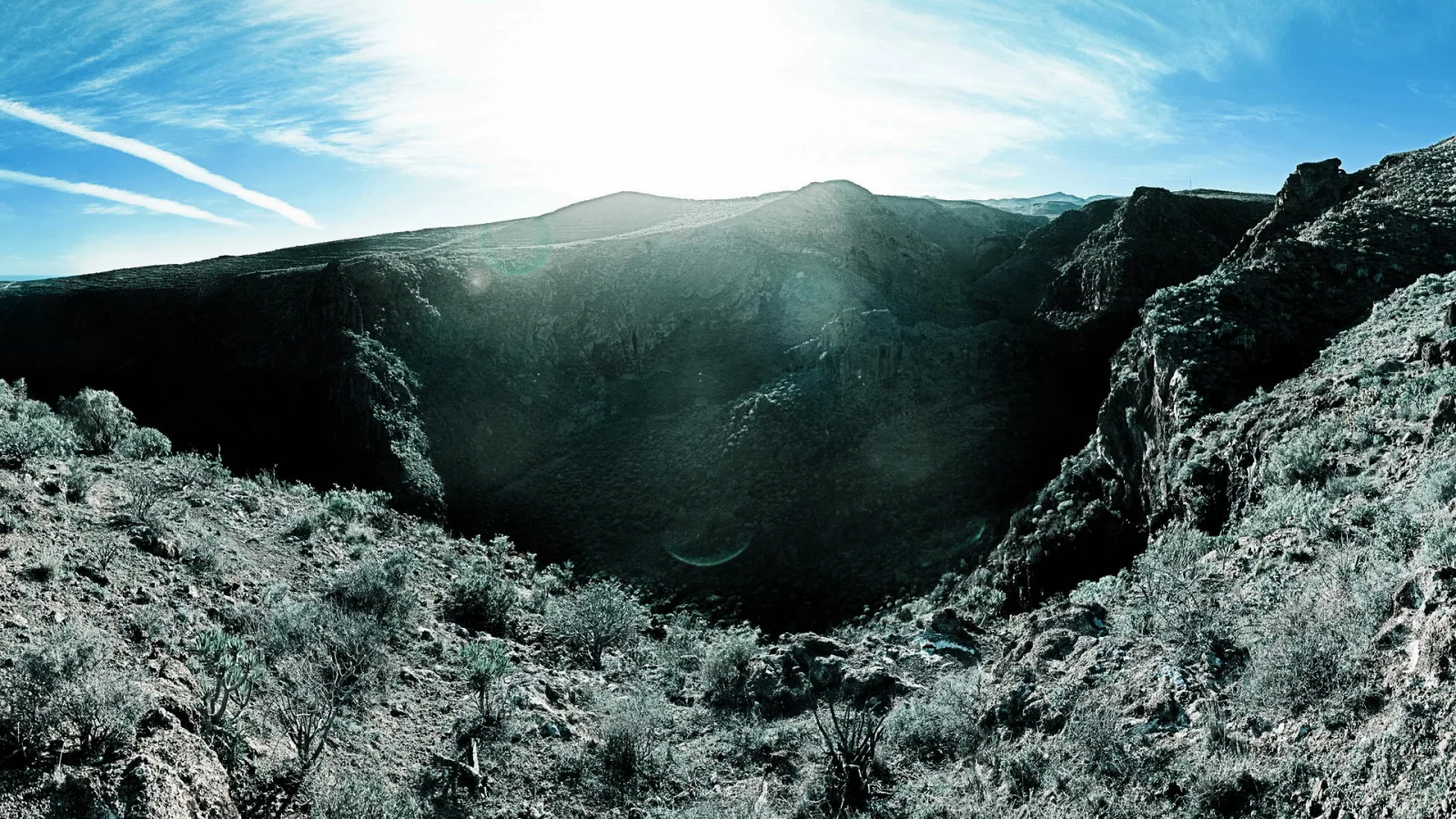Panoramablick in eine tiefe Schlucht mit steilen, dunklen Felswänden, spärlicher Buschvegetation im Vordergrund und strahlender Sonne hinter einem Bergrücken unter blauem Himmel mit Schleierwolken.