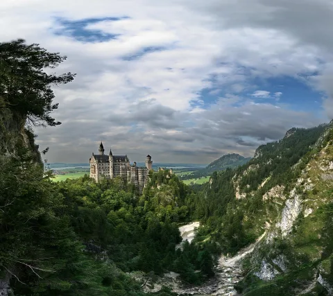 Schloss Neuschwanstein mit hellen Türmen auf einem bewaldeten Felsen, darunter ein Tal mit Fluss, dahinter Berge und eine weite Wiese unter bewölktem Himmel.